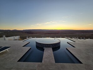 En uendelig pool med et spabad, omgivet af en beton terrasse og bænke, med udsigt over et ørkenlandskab ved solnedgang.