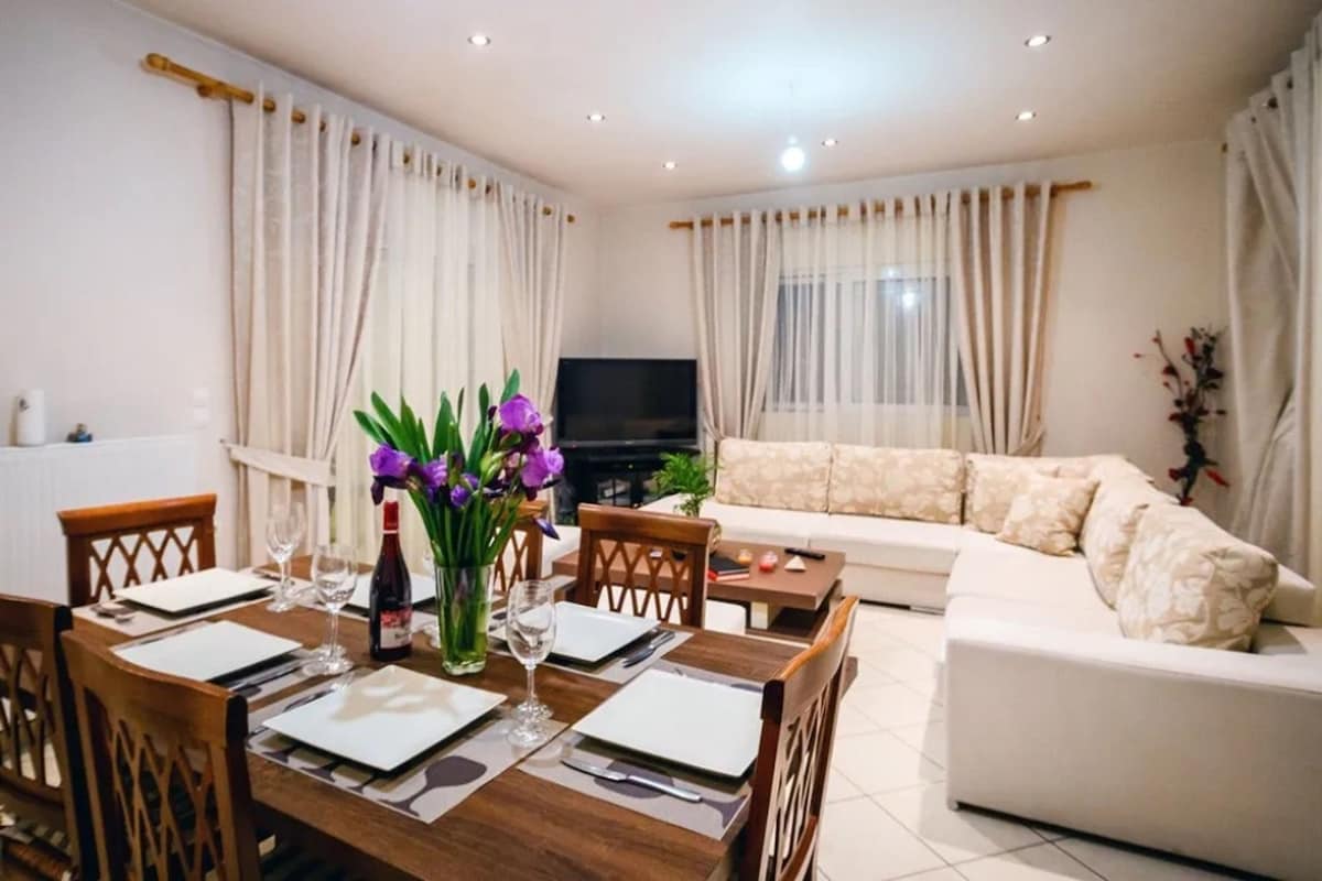 Una sala de estar con una mesa de comedor dispuesta para dos personas, un sofá, un televisor y una planta.