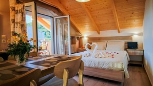 Ein gemütliches Schlafzimmer mit Holzbalkendecke, einem Bett mit weißen Leinen und schwanenförmigen Kissen, einem Nachttisch mit Fernseher sowie einem Essbereich mit Tisch und Stühlen.