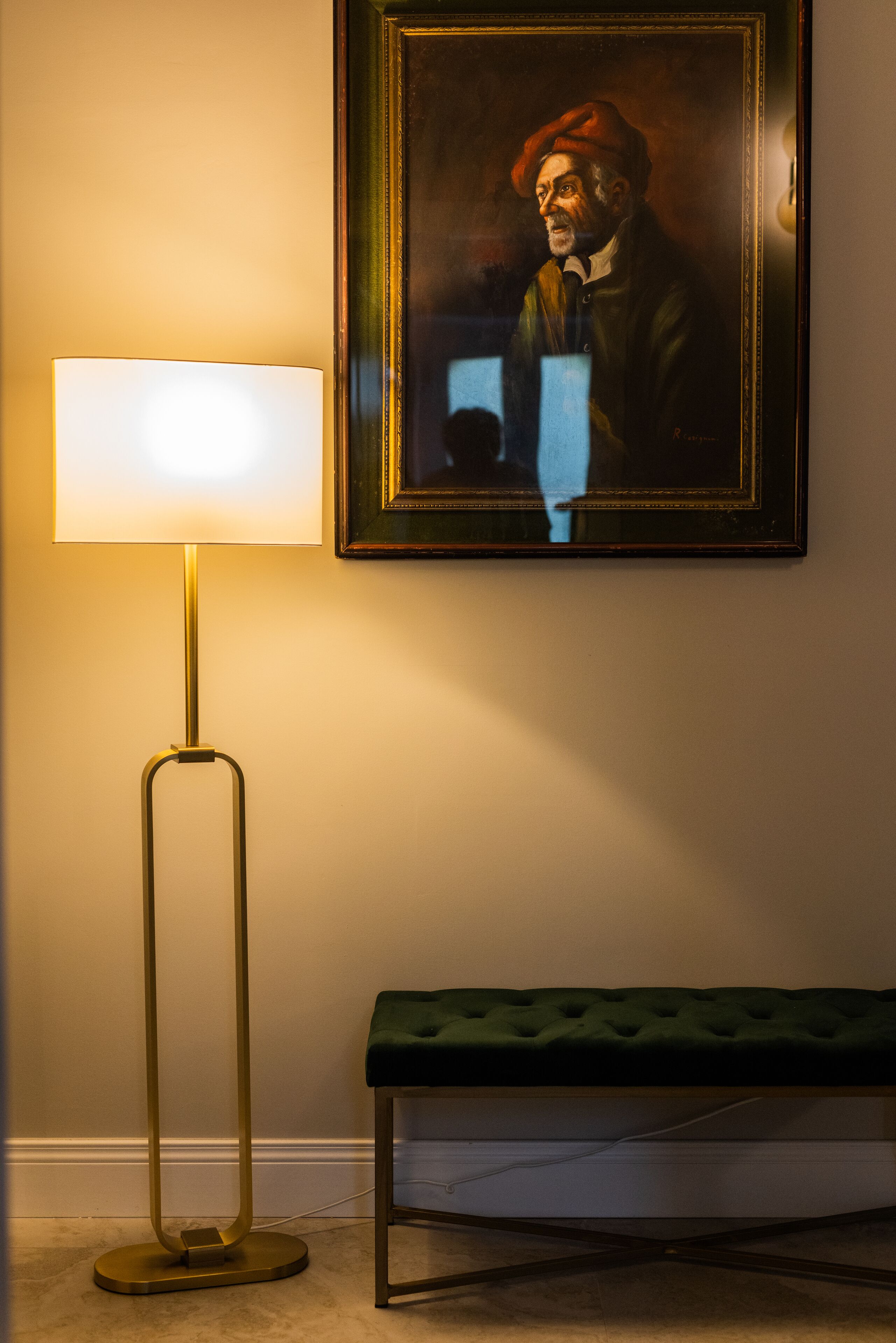 A room with a floor lamp, a framed portrait on the wall, and a green bench.