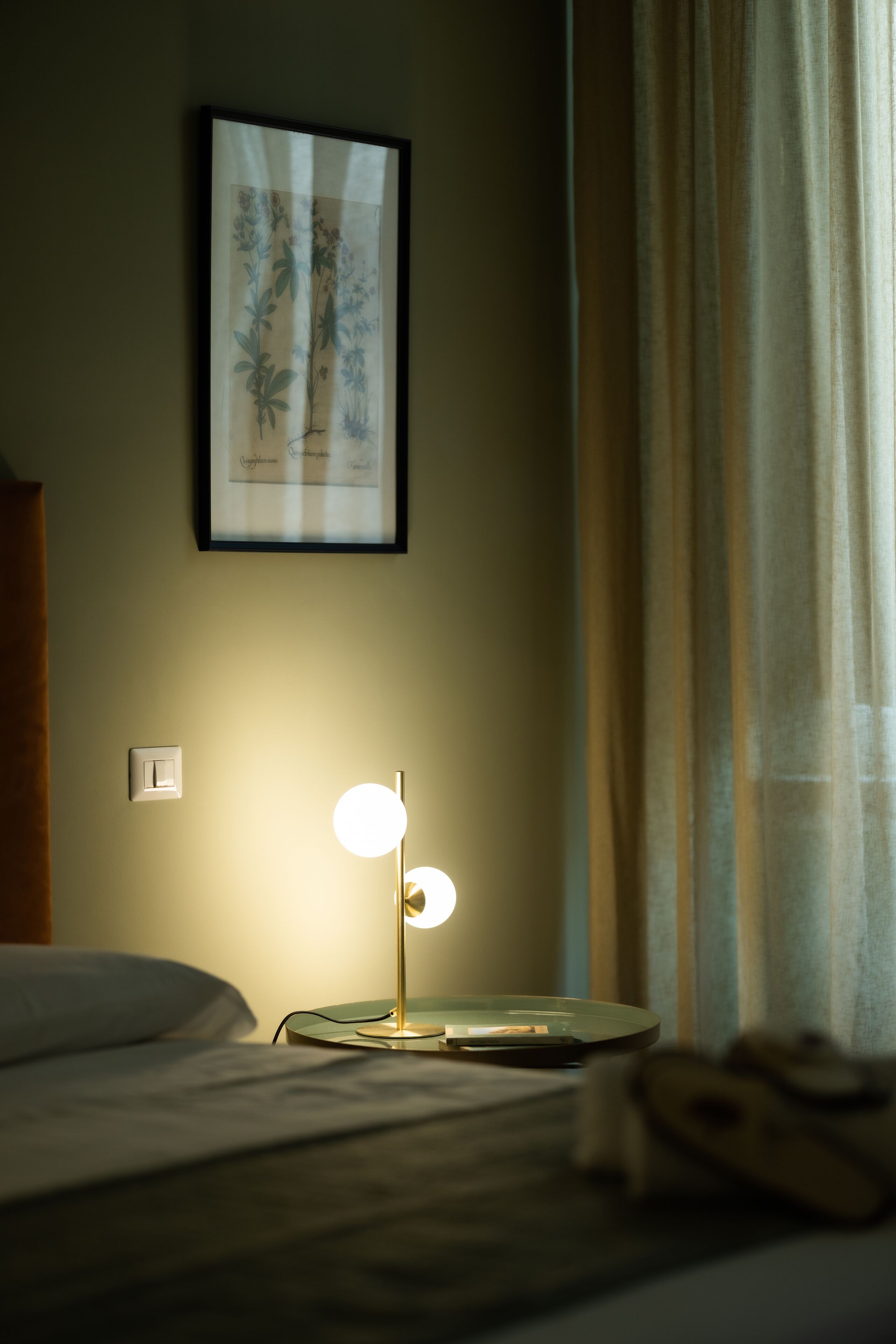 A neatly made bed with a bedside lamp, a framed botanical print on the wall, and a window with curtains.