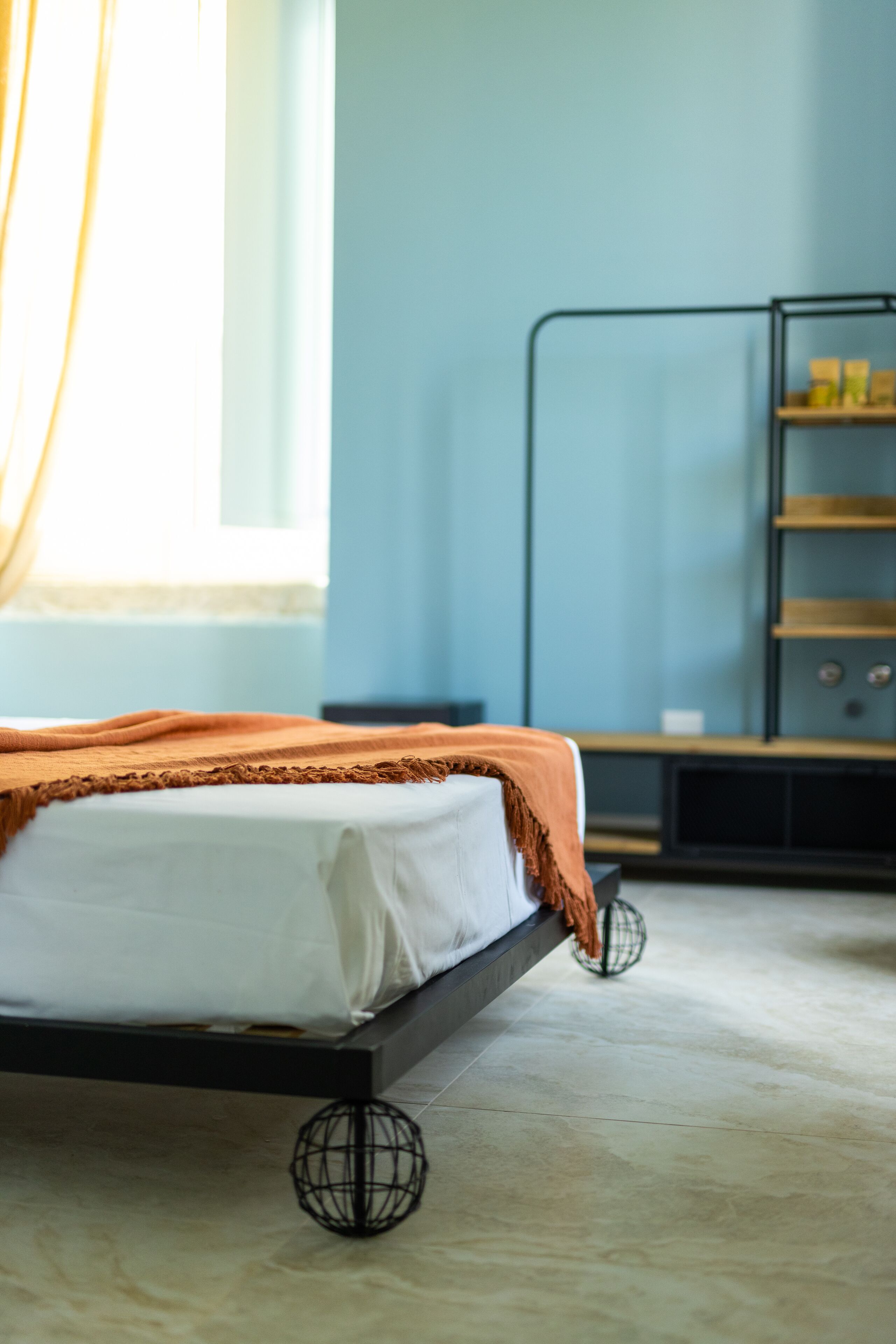 A bed with a white mattress and an orange throw blanket, a black metal shelf unit with books, and a large window with sheer curtains.
