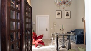 Een eethoek met een ronde tafel, transparante stoelen, een rode stoel, een kroonluchter en lijstwerken aan de muur.