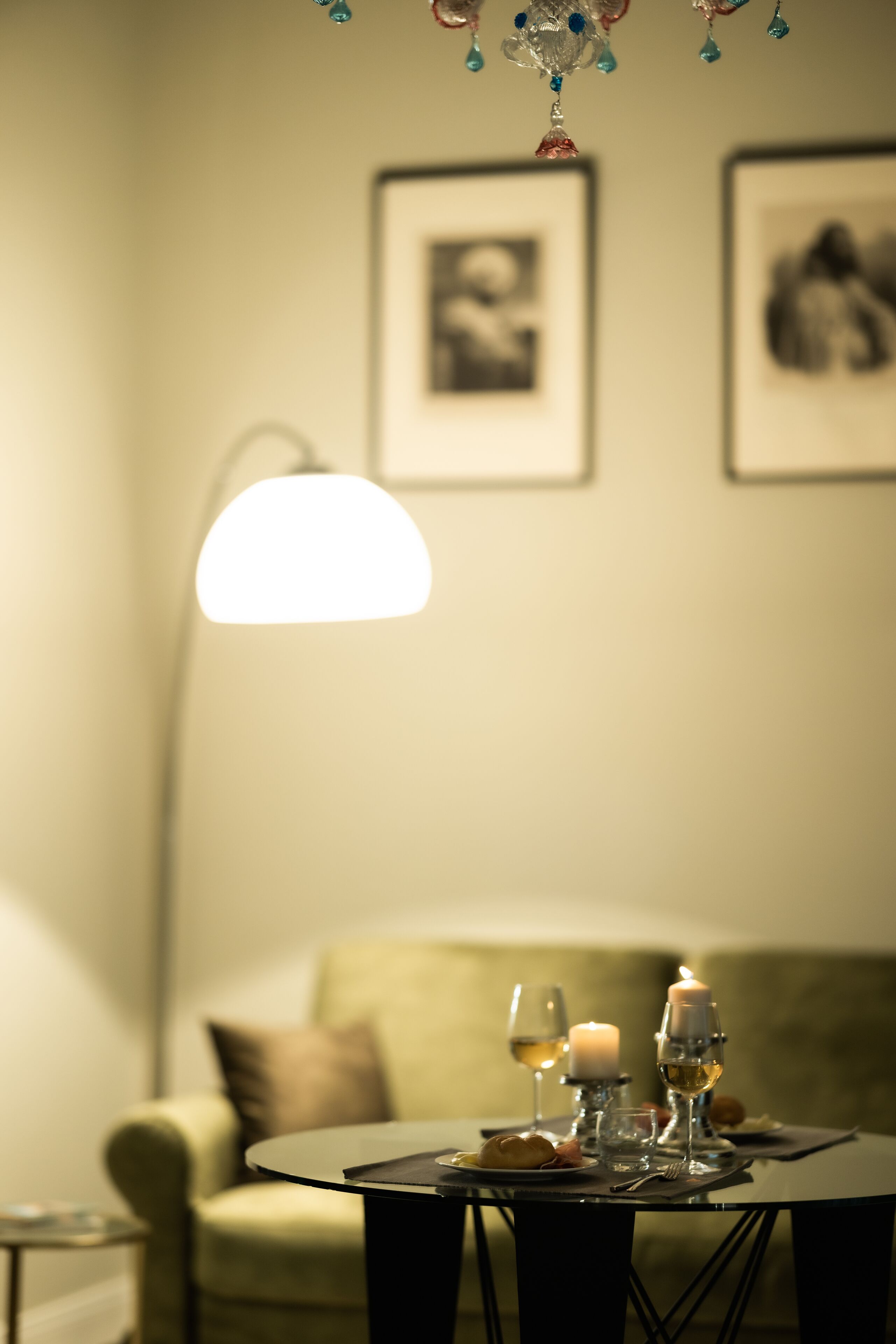 A dining table with a candle, wine glasses, and a plate of food in a softly lit room with a sofa and framed pictures on the wall.