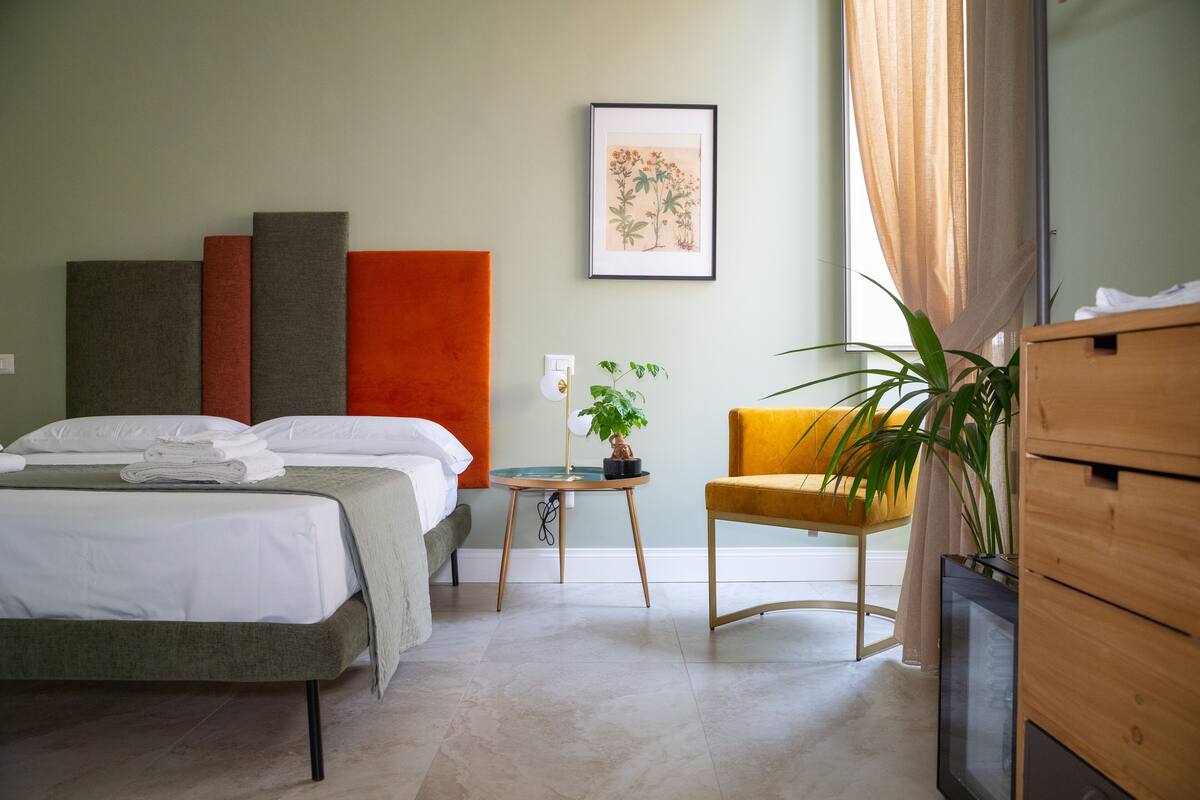 Ein Hotelzimmer mit Bett, Stuhl, einem kleinen Tisch mit Pflanze und einem gerahmten Bild an der Wand.