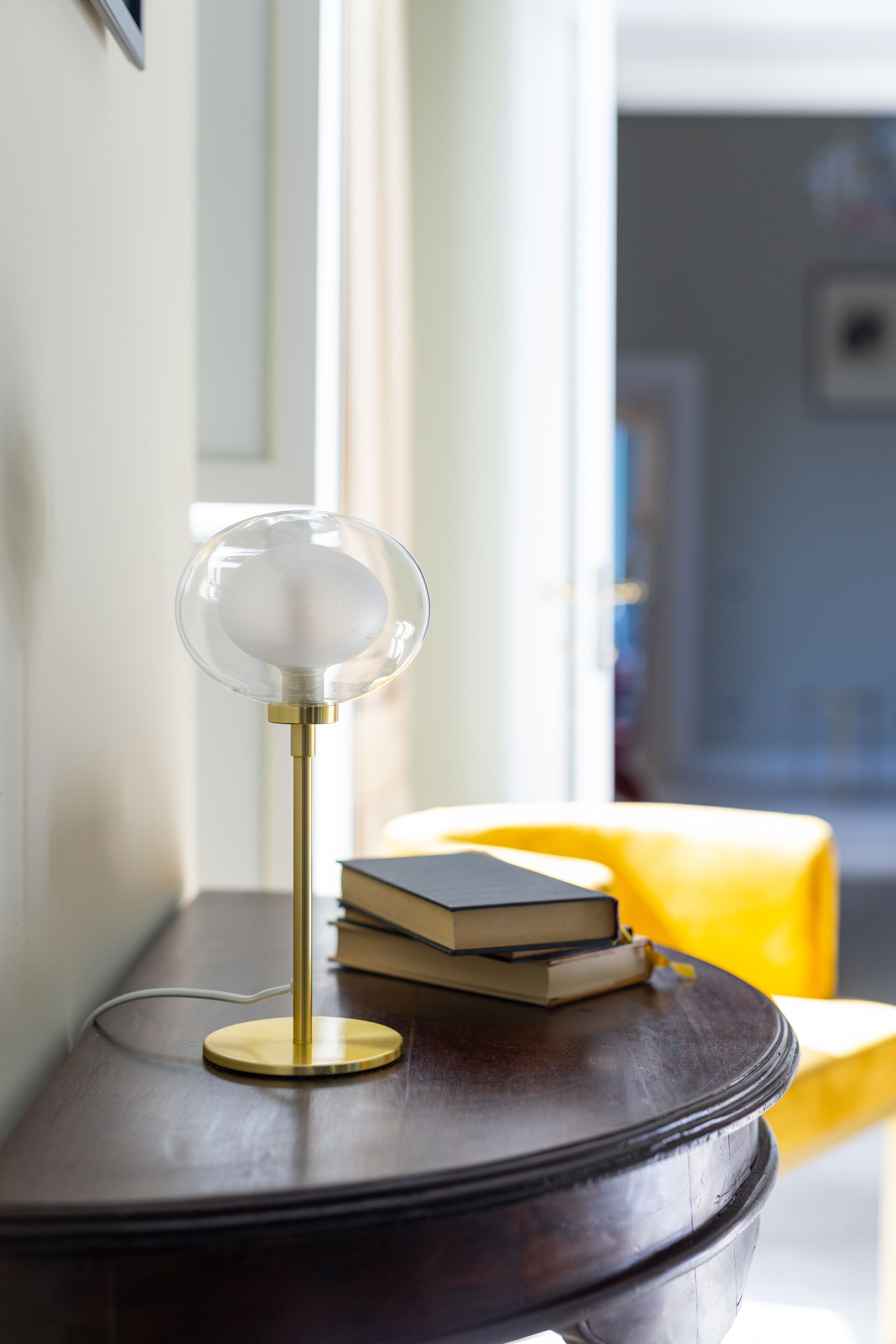 Une table ronde avec une lampe et des livres, une chaise jaune et, au fond, une porte.