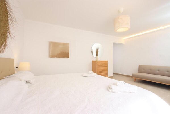 A modern bedroom with a large bed, a bedside table, a mirror, a sofa, and a hanging light fixture.