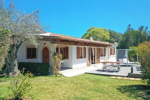 A white villa with a tiled roof, wooden shutters, and a stone pillar. There is a patio with a table and chairs, and a small garden area with a tree and shrubs. - H2927 Finca Alegre near Pollenca for 4 persons with pool (Pollenca)