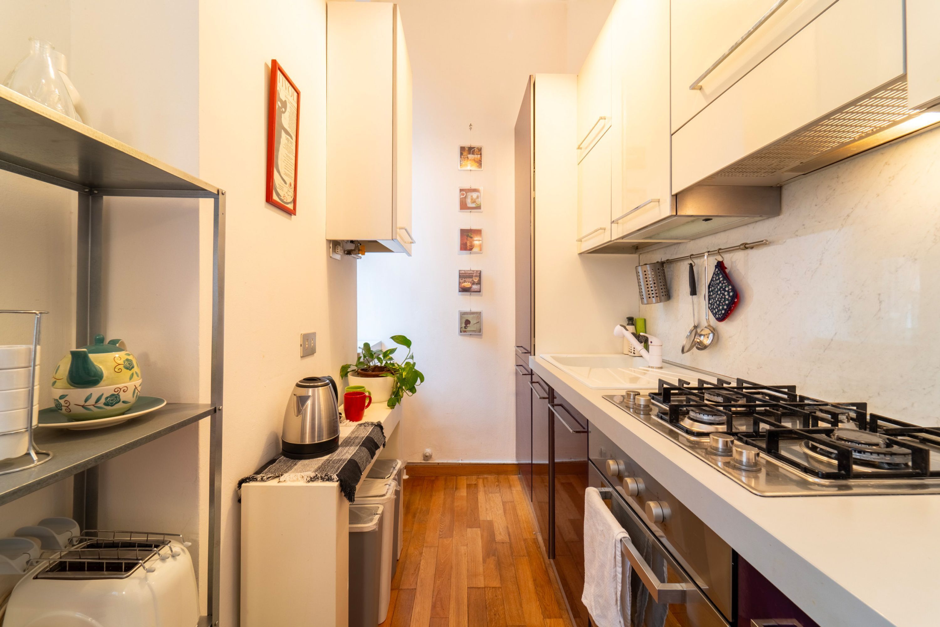 A narrow kitchen with a gas stove, a coffee machine, and a toaster. There are cabinets and a sink on the right side.