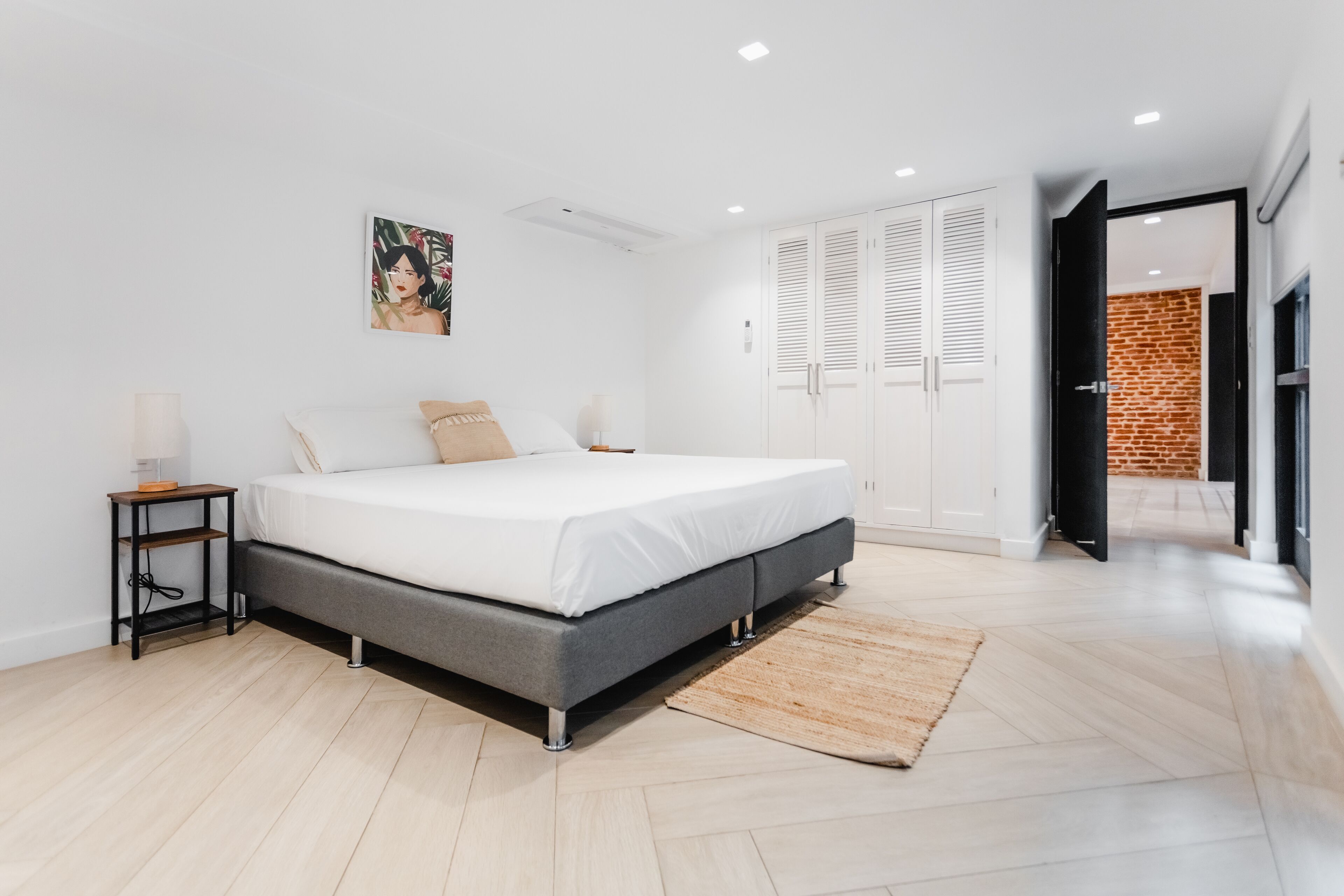 A modern bedroom with a large bed, white walls, and a wooden floor. There is a small bedside table with a lamp, a painting on the wall, and a wardrobe with white doors.