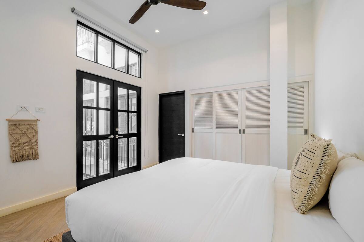 Une chambre avec un grand lit, une porte noire, une fenêtre avec des stores et un ventilateur de plafond.