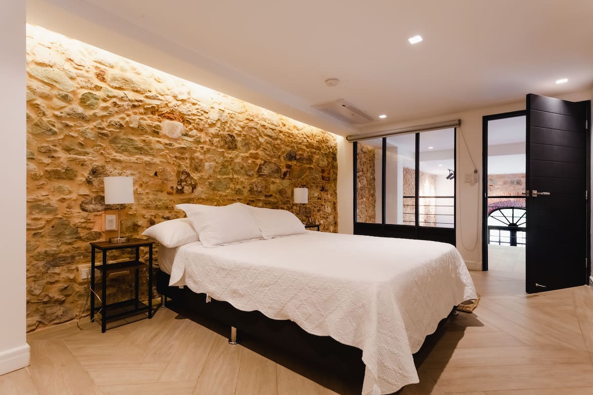 Une chambre avec un mur en pierre, un lit recouvert de draps blancs, une table de chevet avec une lampe, et une vue à travers une porte vitrée.