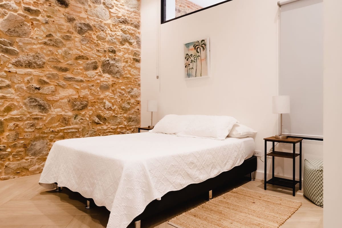 Une chambre avec un mur en pierre, un lit, un ventilateur de plafond, une fenêtre et une table de chevet.