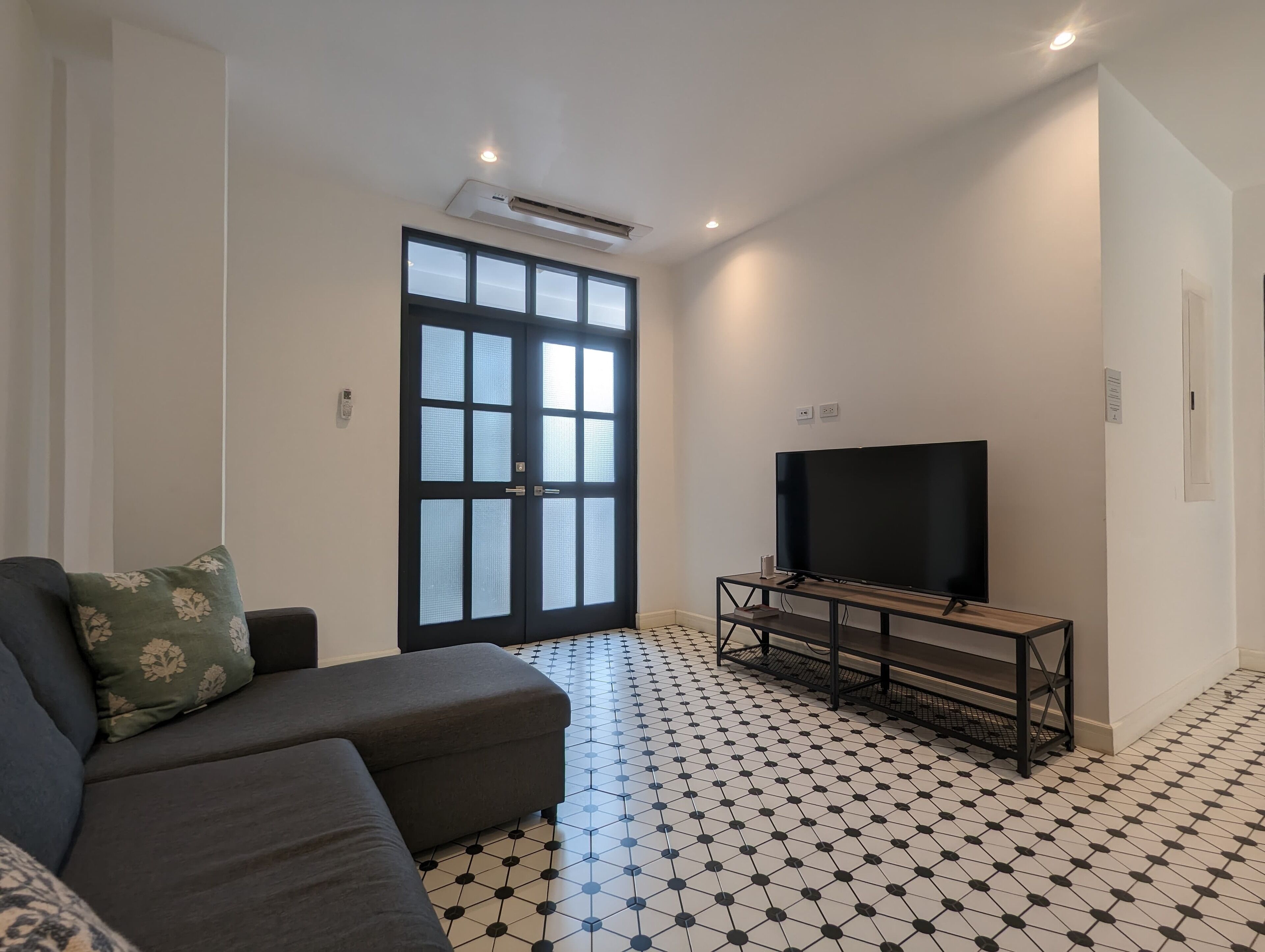 A modern living room with a sectional sofa, a flat-screen TV on a wooden stand, and a large window with blinds.
