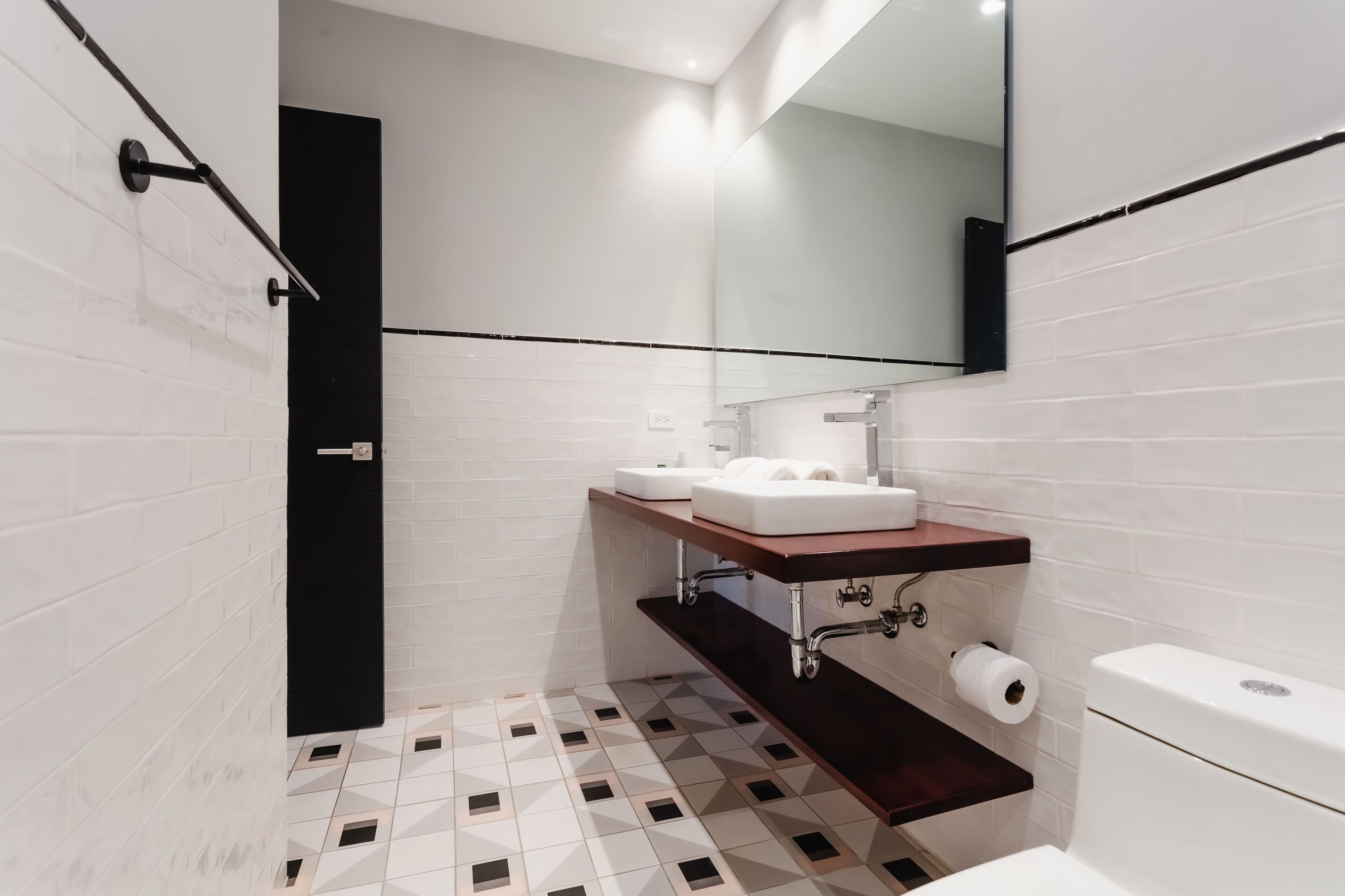 Un baño moderno con piso de baldosas blancas, toallero negro, lavamanos blanco con encimera de madera y un inodoro.