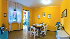 Une pièce aux murs jaunes, une table à manger avec des chaises, un coin cuisine équipé d’appareils électroménagers et une fenêtre avec des rideaux.