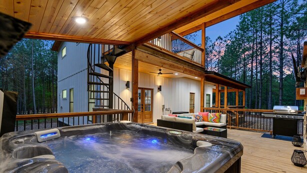 A hot tub area with a wooden deck, a staircase leading to a second floor, and a view of a cabin in the background.