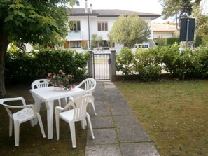 Un patio con una mesa y sillas blancas, rodeado de follaje y una casa al fondo.