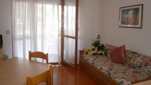 A room with a wooden table and chairs, a sofa with a floral cover, and a door leading to an outdoor area.