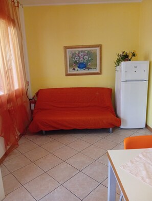 A room with a red sofa, a white refrigerator, a framed floral painting, and a potted plant.