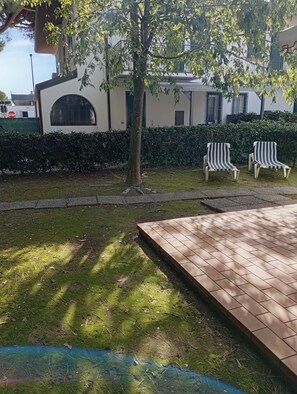 A paved outdoor area with a tree, a hedge, and two striped lounge chairs.