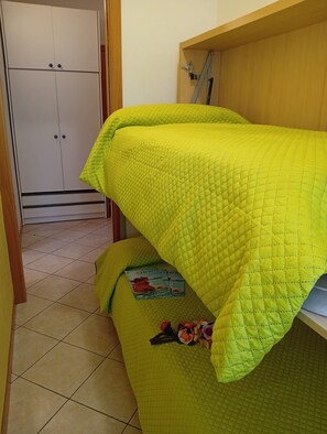 A room with a bed covered in a quilted blanket, a wooden shelf, and a tiled floor.