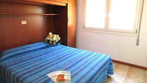 Een slaapkamer met een bed, een raam met zonwering, een houten kast en een kleine tafel met een tijdschrift.