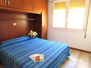 Ein Schlafzimmer mit Bett, einem Fenster mit Jalousien, einem Holzschrank und einem kleinen Tisch mit einer Zeitschrift.