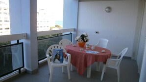 Ein Balkon mit einem Tisch für zwei Personen, weißen Plastikstühlen, einer roten Tischdecke und einer Vase mit Blumen.