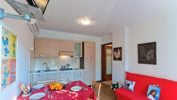 A compact living space with a kitchen, dining area, and a red sofa. - Residenz Elia Wohnung B2 für 5 Personen (Bibione)