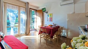 A room with a dining table, chairs, a TV, and a kitchen area. - Residenz Elia Wohnung B2 für 5 Personen (Bibione)