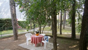 An outdoor seating area with a table and chairs, surrounded by trees and a fence. - Residenz Elia Wohnung Bilo Erdgeschoss für 5 Personen (Bibione)