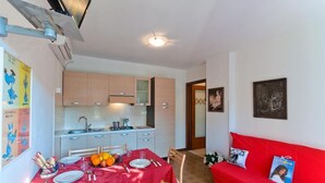 A compact living space with a dining area, kitchenette, and a red sofa. - Residenz Elia Wohnung Bilo Erdgeschoss für 5 Personen (Bibione)