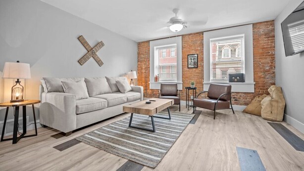 A modern living room with a sofa, coffee table, and chairs, a brick accent wall, and a ceiling fan. - Rhythm Loft Above the Honky Tonk (Saint Charles)