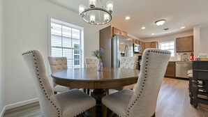 A dining area with a round table and four upholstered chairs, a chandelier, and a kitchen in the background.