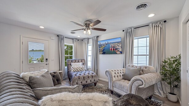 A spacious living room with a large sofa, a tufted armchair, and a ceiling fan. There is a large window with curtains, a door leading outside, and a potted plant.