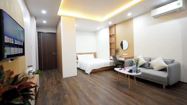 A modern hotel room with a bed, a grey sofa, a small table, a TV, and a desk. - Fusion Gates Apartment (Hanoi)
