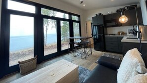 A modern kitchen with dark cabinets, a dining area with a table and chairs, and large windows offering a view of the sea. - Crisp Point Beach Cabin (Newberry)