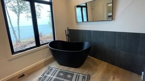 A modern bathroom with a freestanding bathtub, a large window with a view of trees and water, and a mirror on the wall. - Crisp Point Beach Cabin (Newberry)