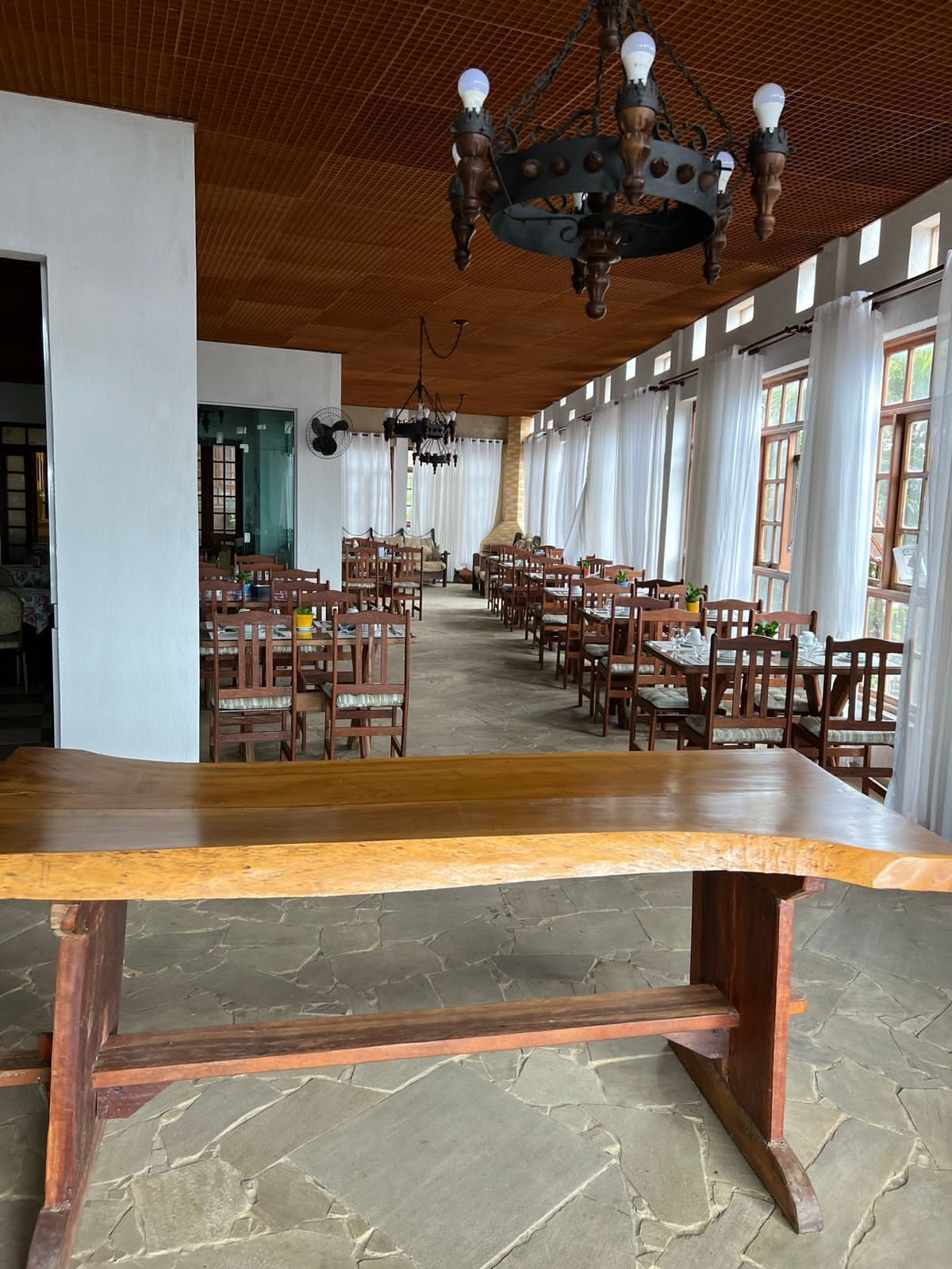 Una àmplia sala de dinar amb una llarga taula de fusta en primer pla, envoltada per diverses taules i cadiretes. Un gran candelabrum penja del sostre, i la sala té un sostre alt amb acabat de fusta.