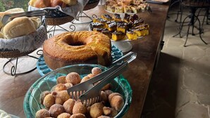 Una mesa de buffet con diversas opciones de panadería y repostería, incluyendo pan, pasteles y rosquillas, en un espacio con vistas a edificios y zonas verdes.