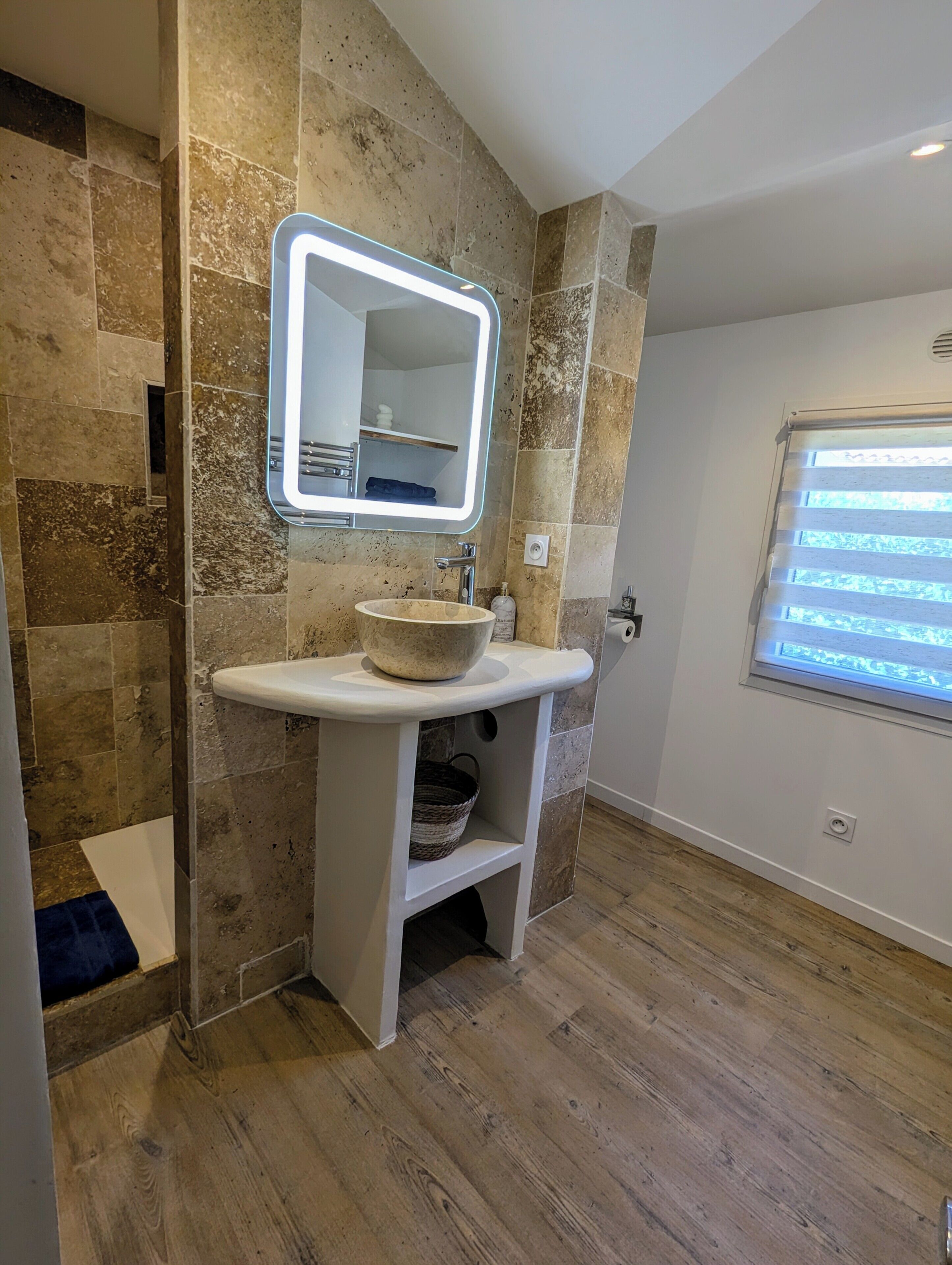 Een badkamer met een stenen muur, een wit wastafelmeubel met een stenen kom, een spiegel met verlichting, en een raam met jaloezieën.