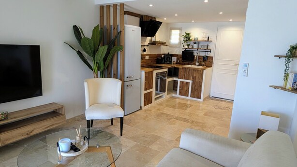 Ein modernes Wohnzimmer mit einem beigen Sofa, einem Glastisch und einem Fernseher. Im Hintergrund ist eine Küche mit Holzschränken und einem weißen Kühlschrank erkennbar.
