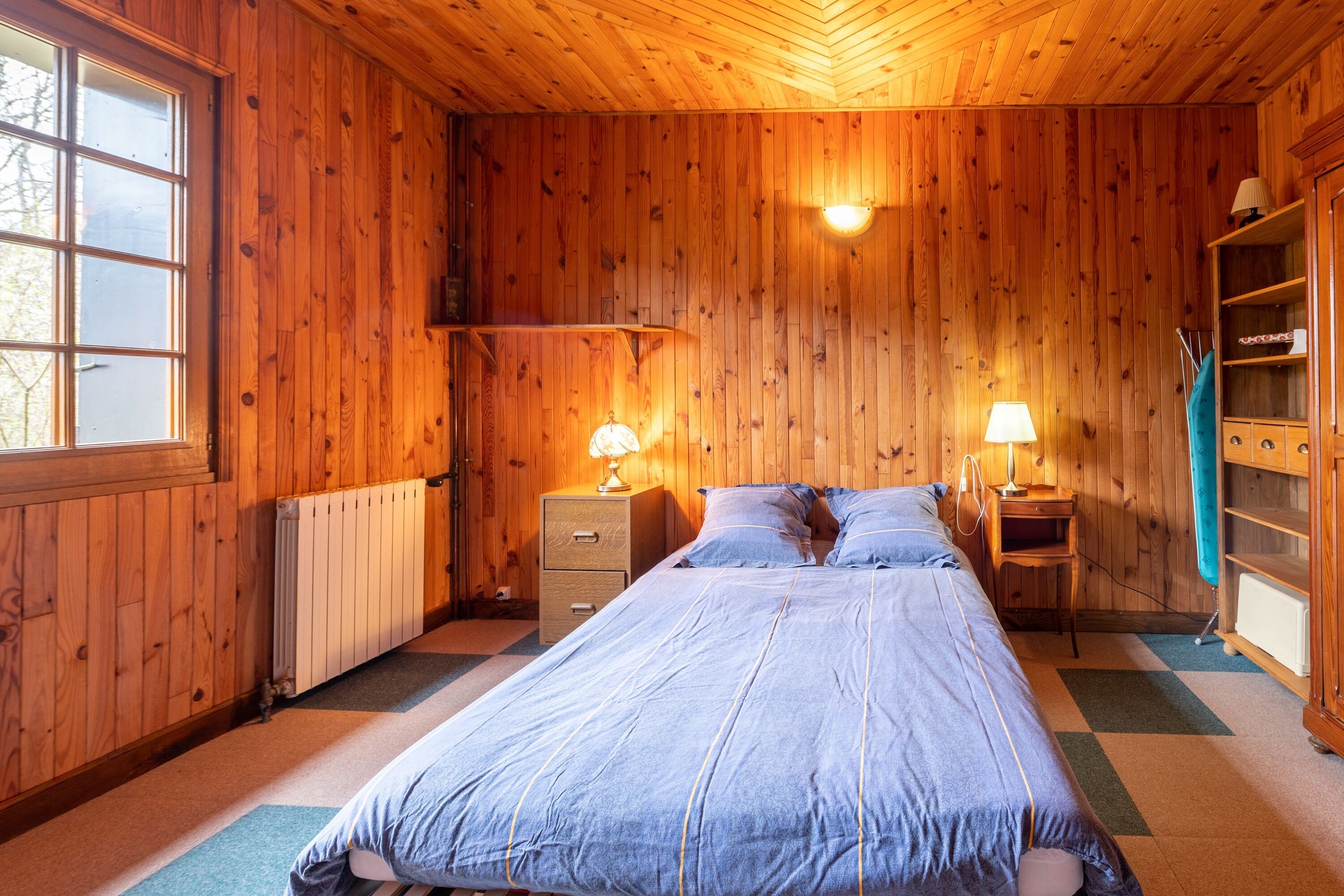 A wooden-paneled bedroom with a bed, two bedside tables with lamps, a radiator, and a wardrobe.