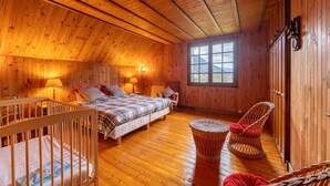 Ein gemütliches Holzhütten-Zimmer mit schrägem Dach, einem großen Bett mit Steppdecke, zwei Stühlen, einem Babybett und einem Fenster mit Blick auf die Berge.