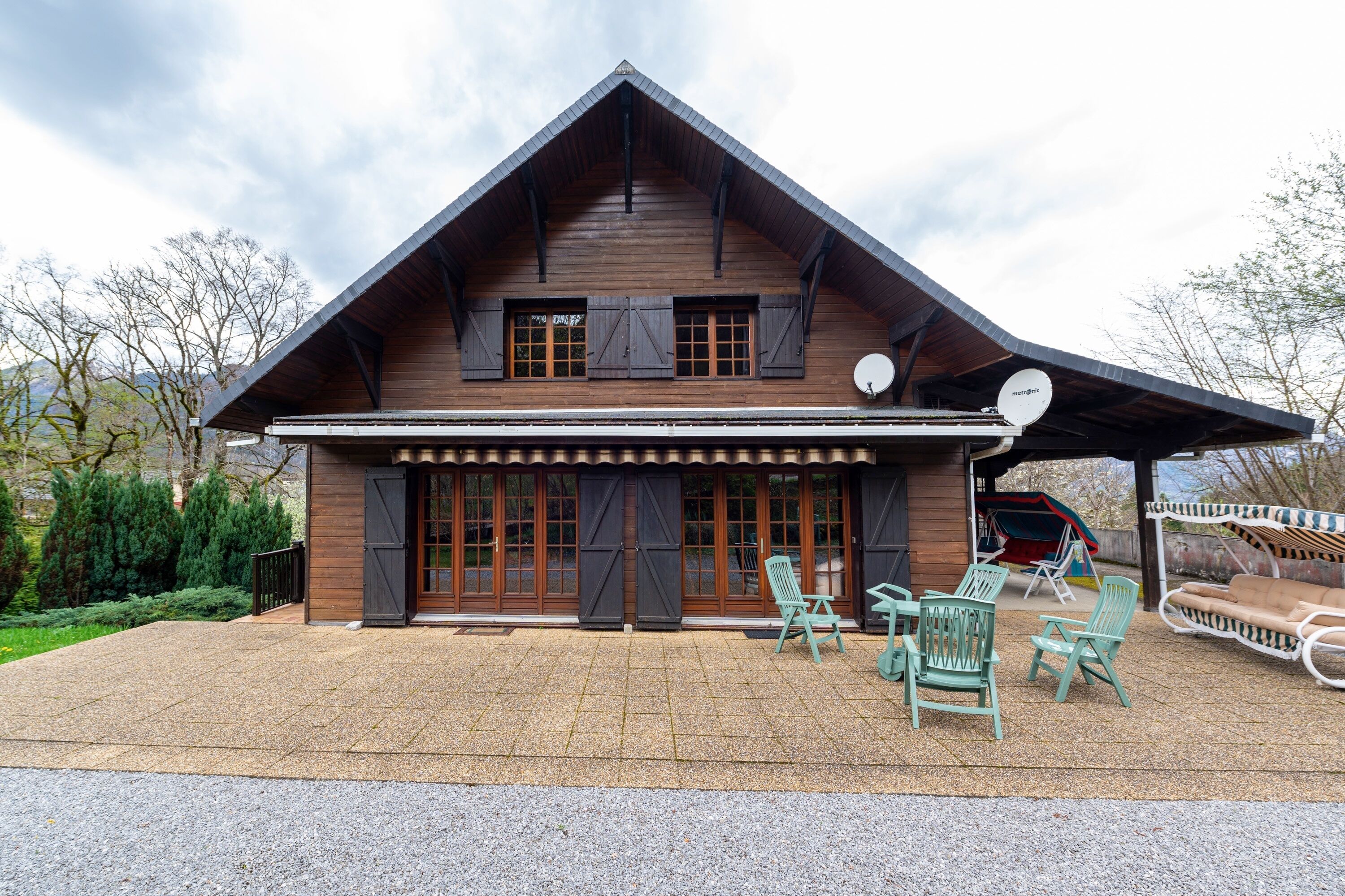 Een houten chalet met een schuin dak, grote ramen en een overdekt terras met buitenmeubilair.