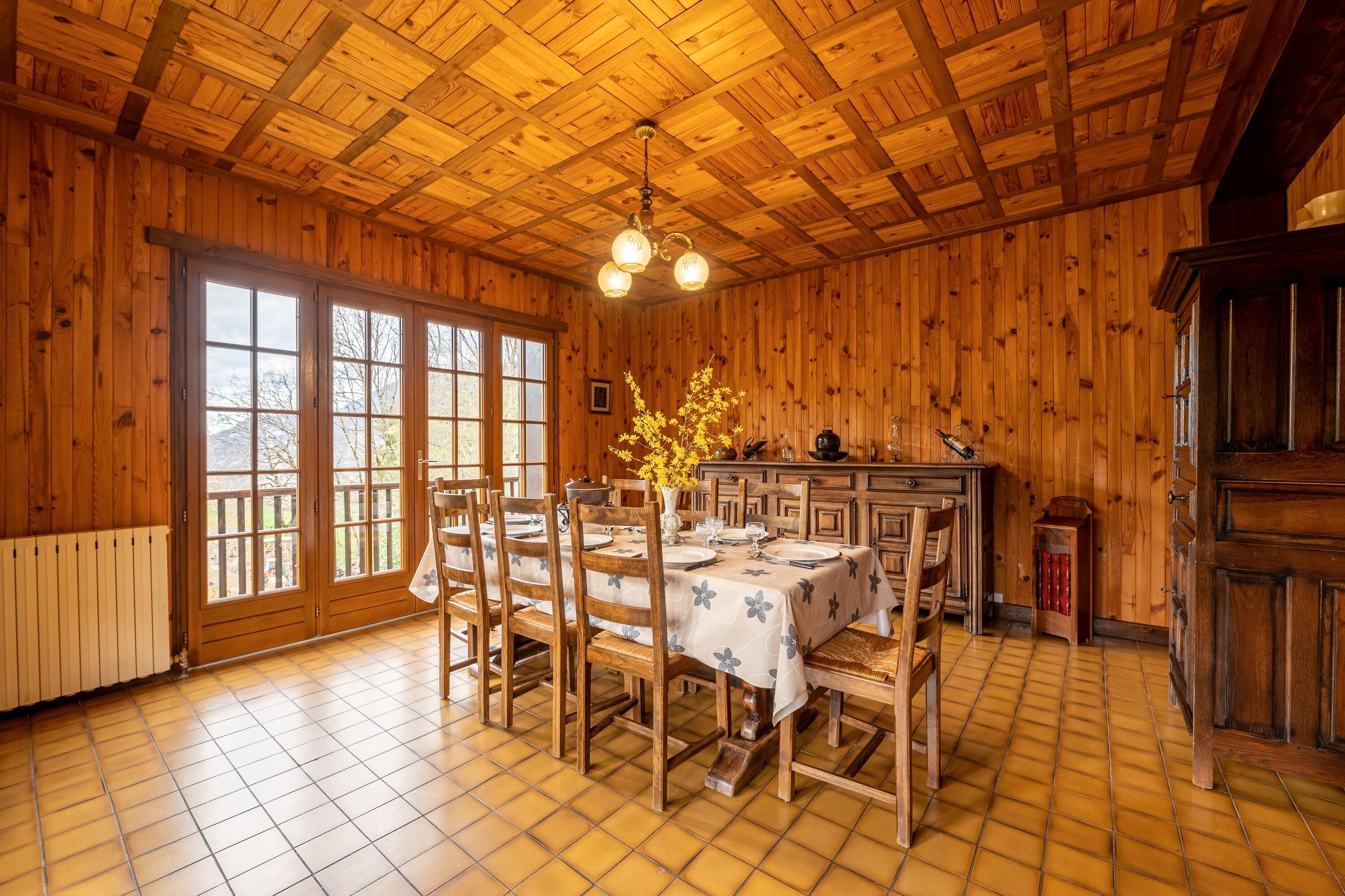 A wooden interior with a dining table set for four, a kitchen area, and a large window with a view of trees.