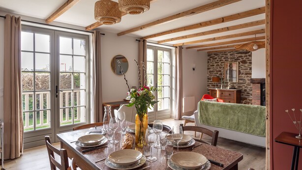 Un coin repas avec une table en bois dressée pour quatre personnes, un vase rempli de fleurs et un mur en pierre en arrière-plan.