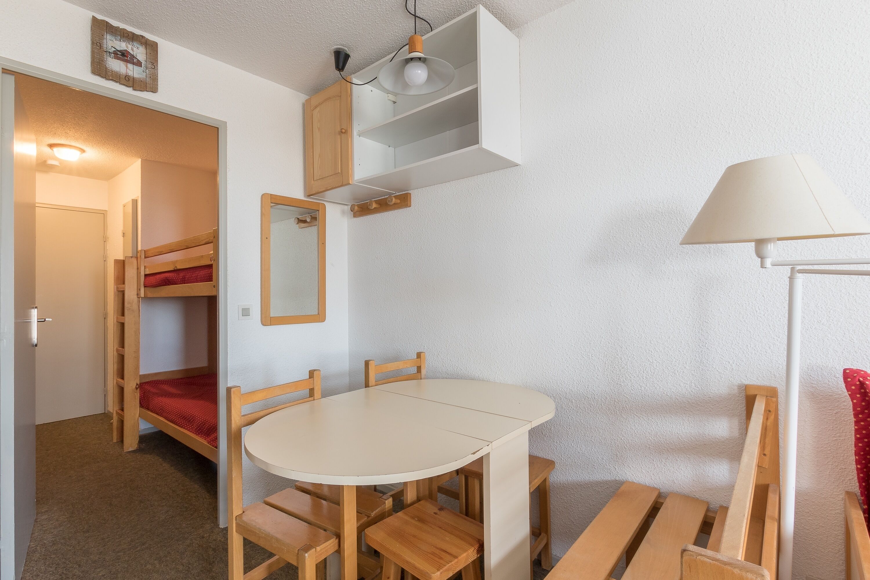 A compact room with a bunk bed, a round table with chairs, a lamp, and a shelf with a mirror.