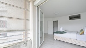 A modern bedroom with a sliding glass door leading to a balcony, a bed with a tufted headboard, and a small built-in shelf above the bed.